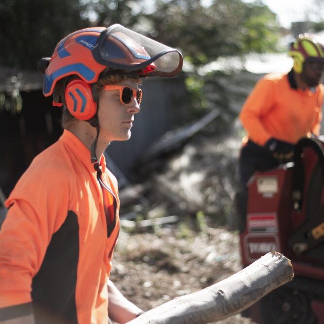 Two men wearing orange safety gear and holding a log.